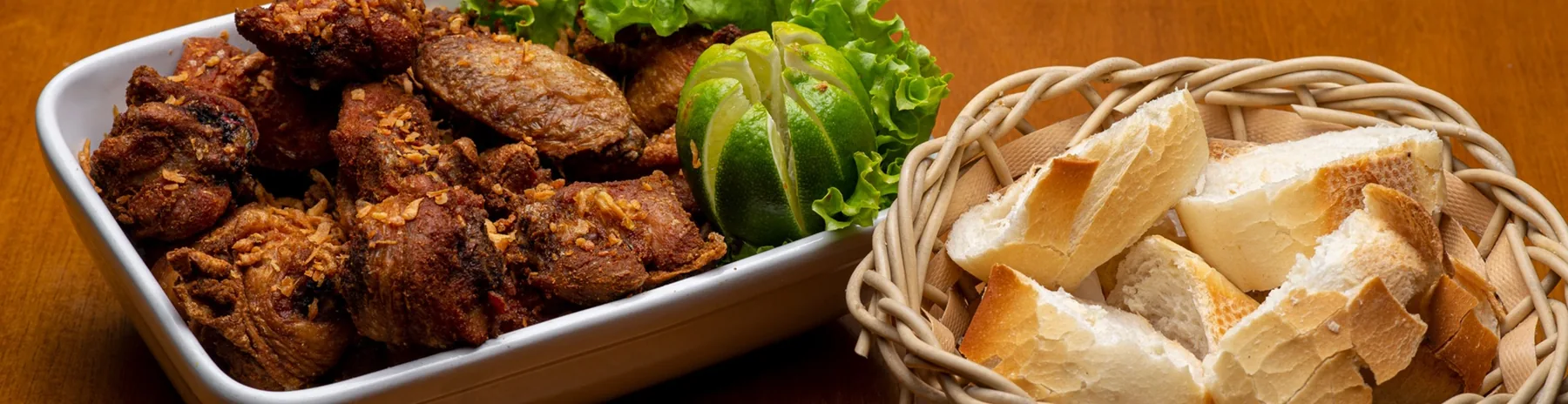 Porção de frango a passarinho com alho frito, limão e alface, ao lado de uma cesta de pães.