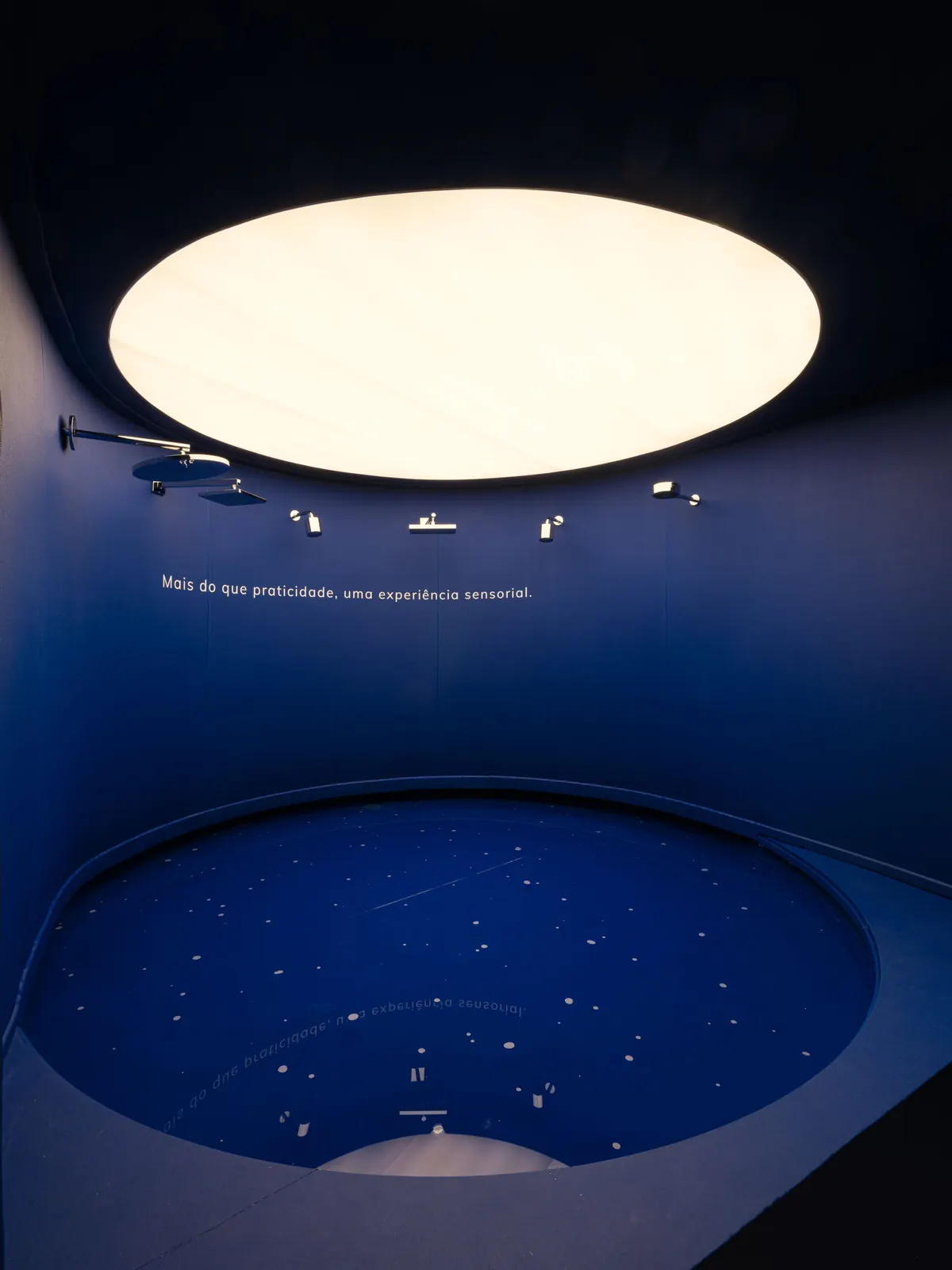 Área imersiva do estande: sala circular escura com um grande óculo de luz no teto, chuveiros na parede e a frase “Mais do que praticidade, uma experiência sensorial.”.
