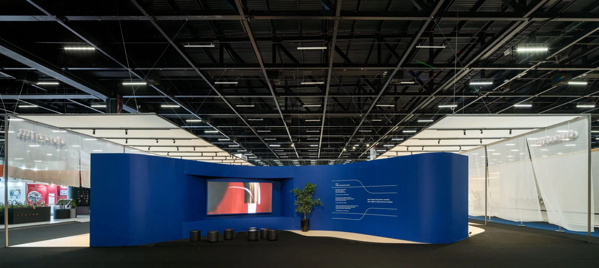 Vista panorâmica do estande: parede azul ondulada com tela de LED ao centro, texto manifesto, bancos e cortinas translúcidas dos dois lados.