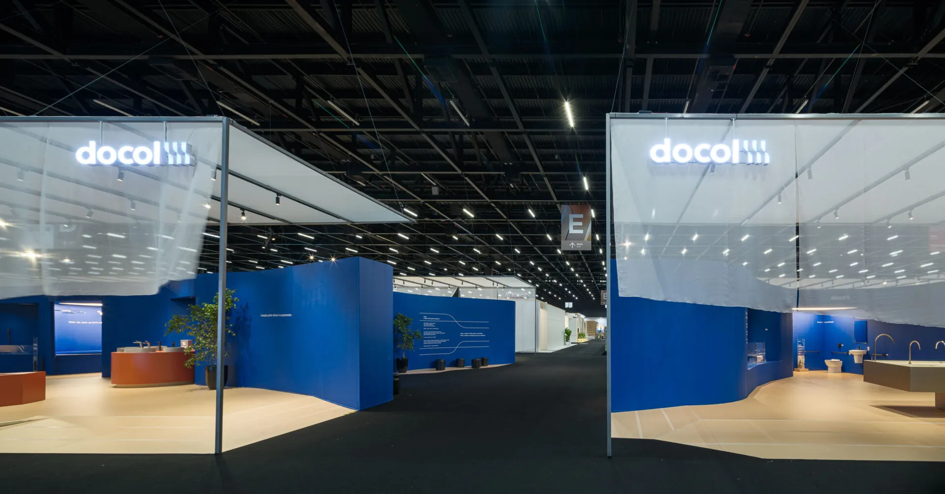 Entrada do estande da Docol na Expo Revestir 2026: duas caixas de cortina translúcida com o logo docol iluminado, frente a frente, e as paredes azuis curvas ao fundo.