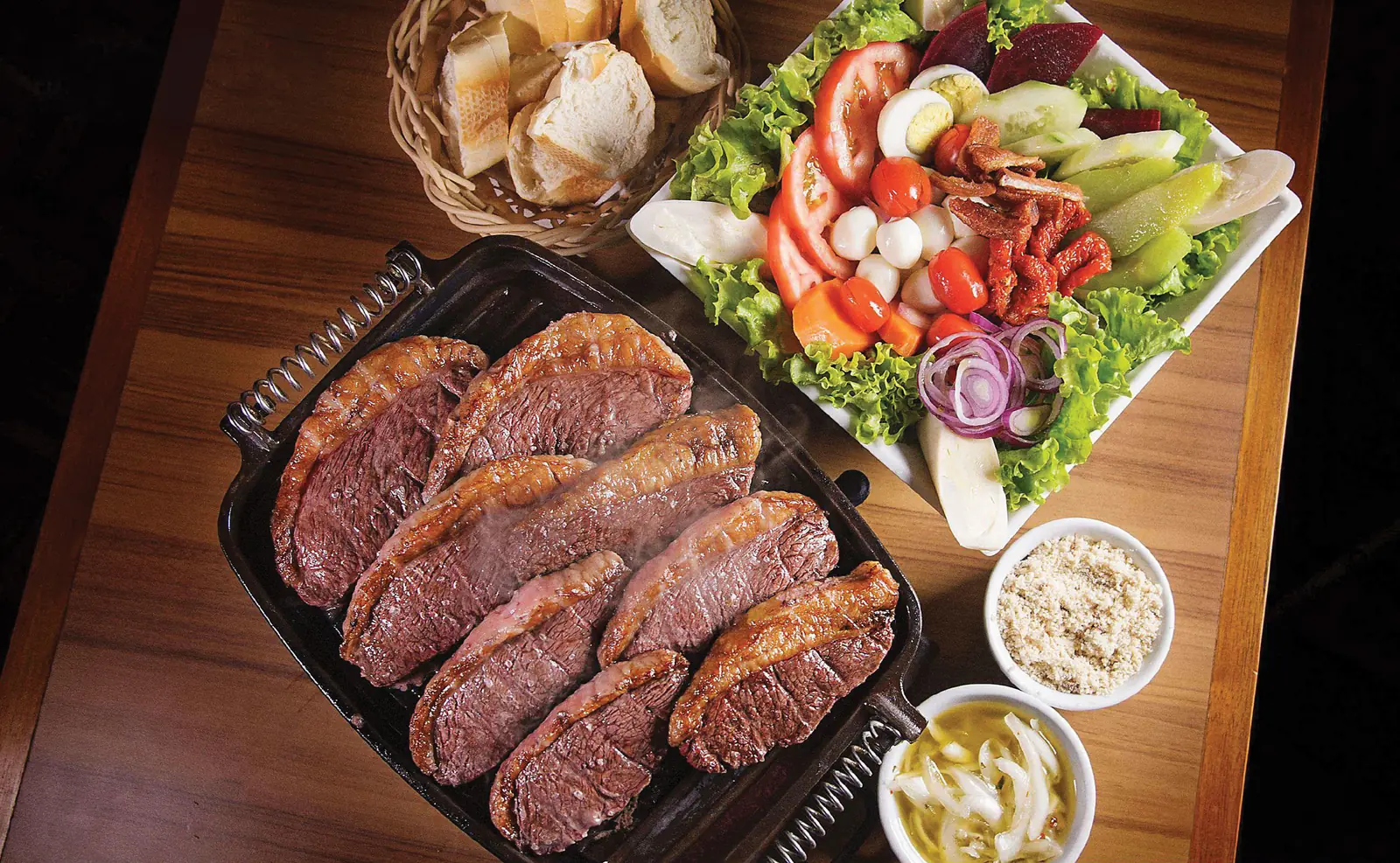 Choupana: picanha fatiada na chapa, com salada, pão, farofa e vinagrete sobre mesa de madeira.