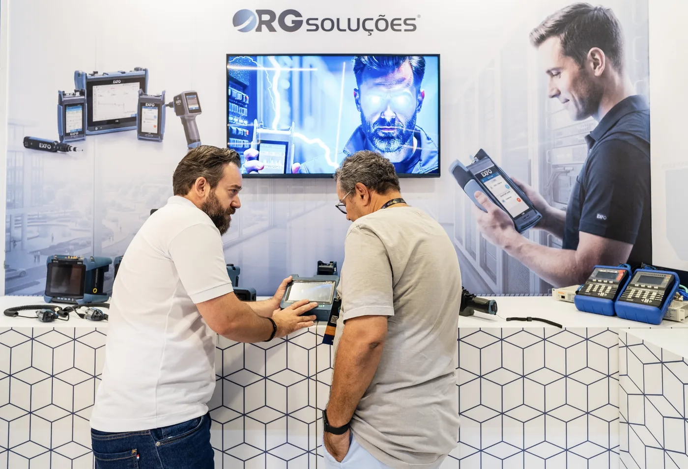 Demonstração de equipamento no estande da RG Soluções, diante do painel com produtos e a campanha Fiber Hero.