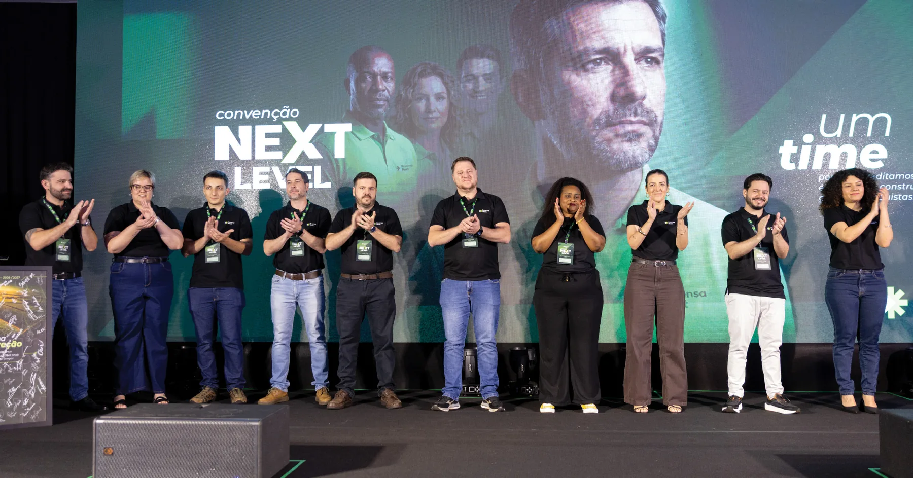 Equipe aplaudindo no palco da convenção Next Level, com o painel “convenção NEXT LEVEL · um time”.